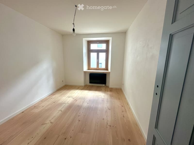 Empty room with wooden flooring, white walls, and a small window with a radiator below it.