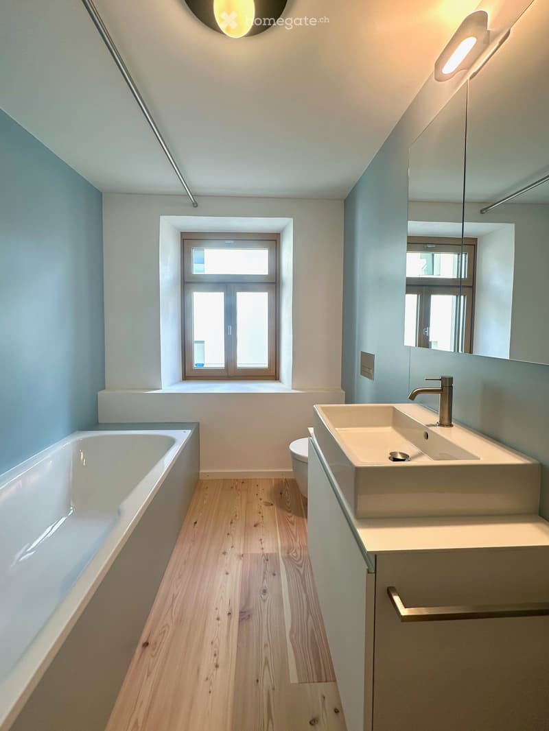 Modern bathroom with a bathtub, sink, toilet, and large mirror. Light blue walls, wooden flooring, and a window providing natural light.