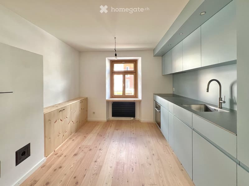 Modern kitchen with light grey cabinets, wooden flooring, and a stainless steel sink with a modern faucet. The space features built-in cabinets and a window with a radiator below.