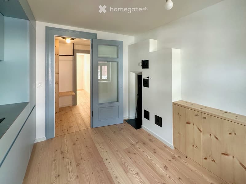 Empty hallway with light wooden flooring, white walls, and a grey door leading to another room. There is a small wooden bench visible in the adjacent room.