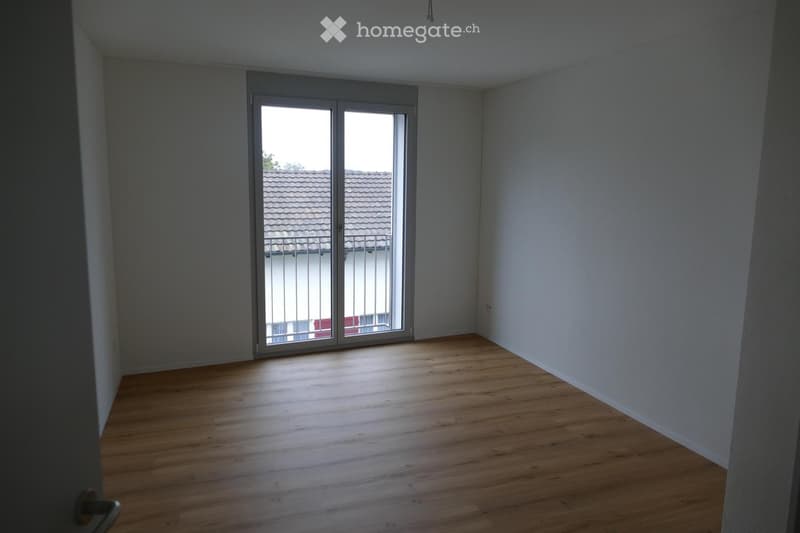 Empty room with wooden flooring and large sliding glass doors leading to a balcony. The walls are painted in neutral tones, and the space is well-lit with natural light.