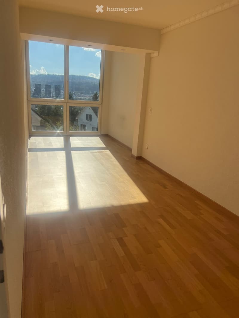 Empty room with wooden flooring and large windows offering a scenic view of mountains and cityscape.