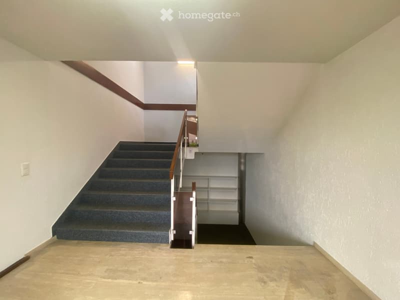 A modern hallway with a staircase leading upwards, featuring a wooden handrail, gray carpeted stairs, and white walls.