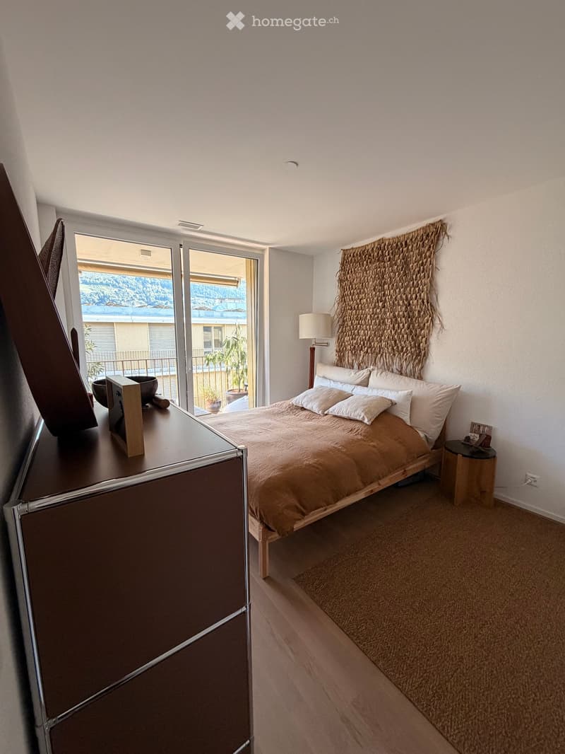 Modern bedroom with a large bed, nightstand, table, and a view of the balcony and surrounding area.
