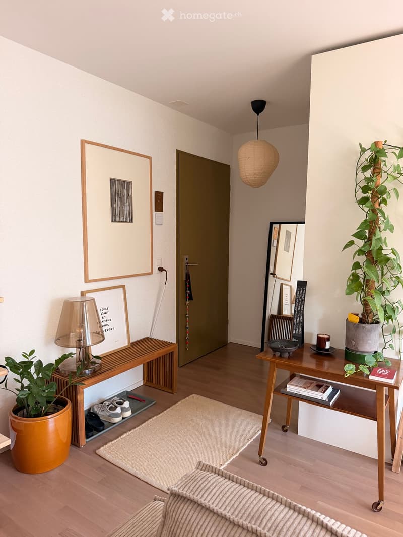 A modern, minimalist living room with light wooden flooring, a beige sofa, a small wooden table with a lamp, a potted plant, and several picture frames on the walls.