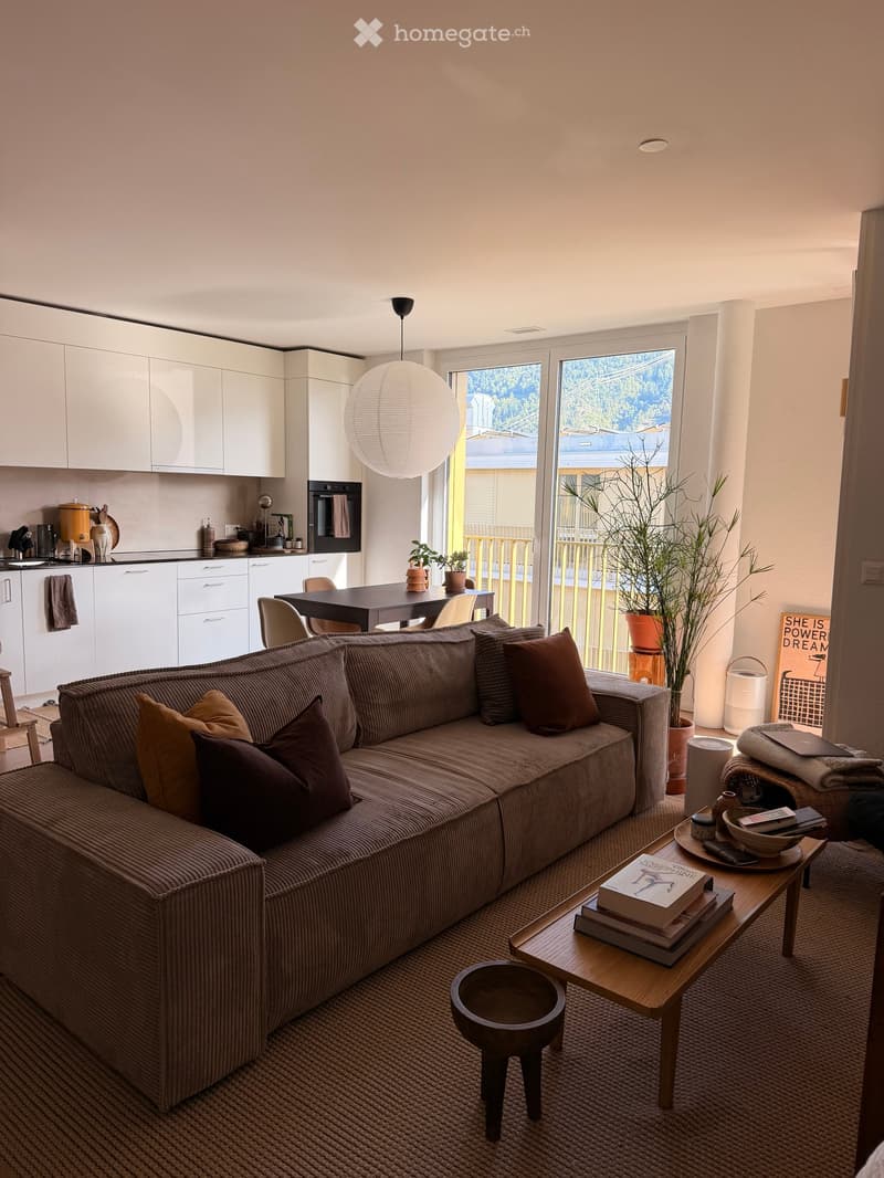 Modern living room with a sectional sofa, coffee table, and kitchen area visible. Features large windows with a balcony view, potted plants, and decorative items.