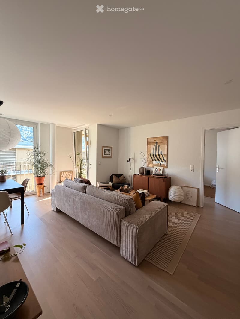 Modern living room with light wood flooring, large window, balcony access, sofa, coffee table, side chair, dining table, cabinet, and decorative items.