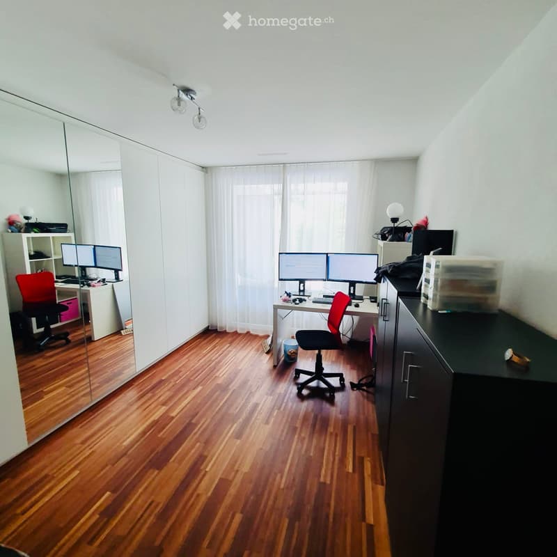 A modern, well-lit room with wooden flooring, two desks, multiple screens, and a large mirror on the wall.
