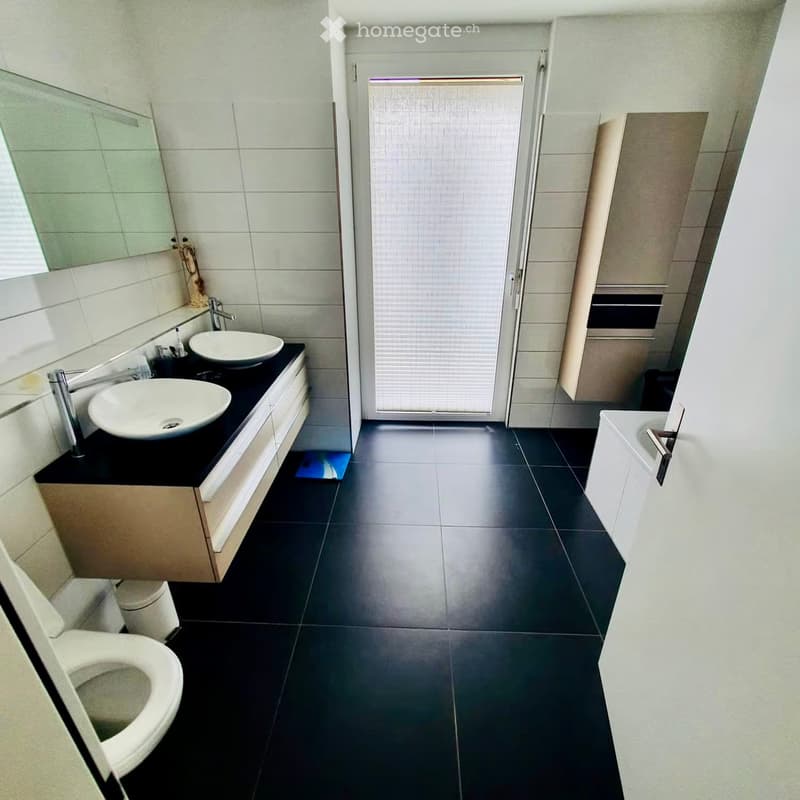 Modern bathroom with black tiled floor, white sink, mirror, toilet, and bathtub. Features a sleek, contemporary design with a glass shower enclosure.