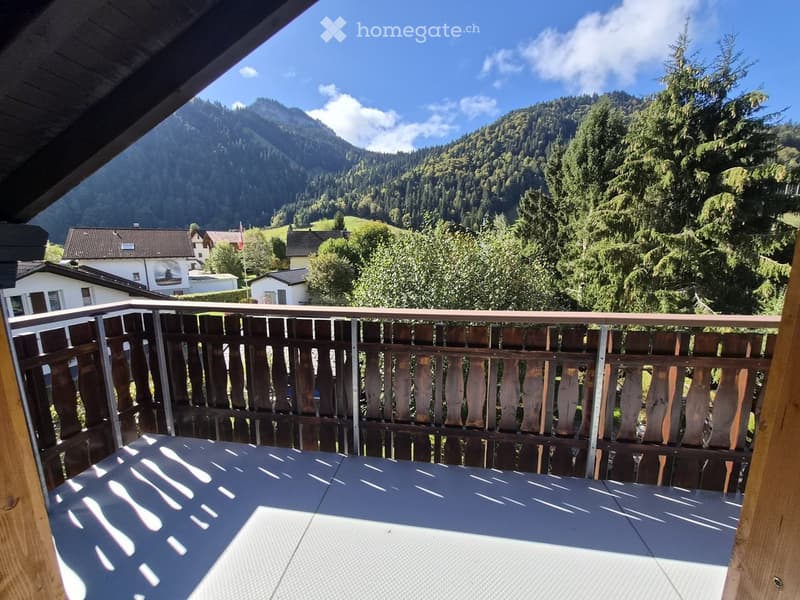 Balcony with wooden railing, overlooking a scenic mountain landscape and forested area with houses in the distance.