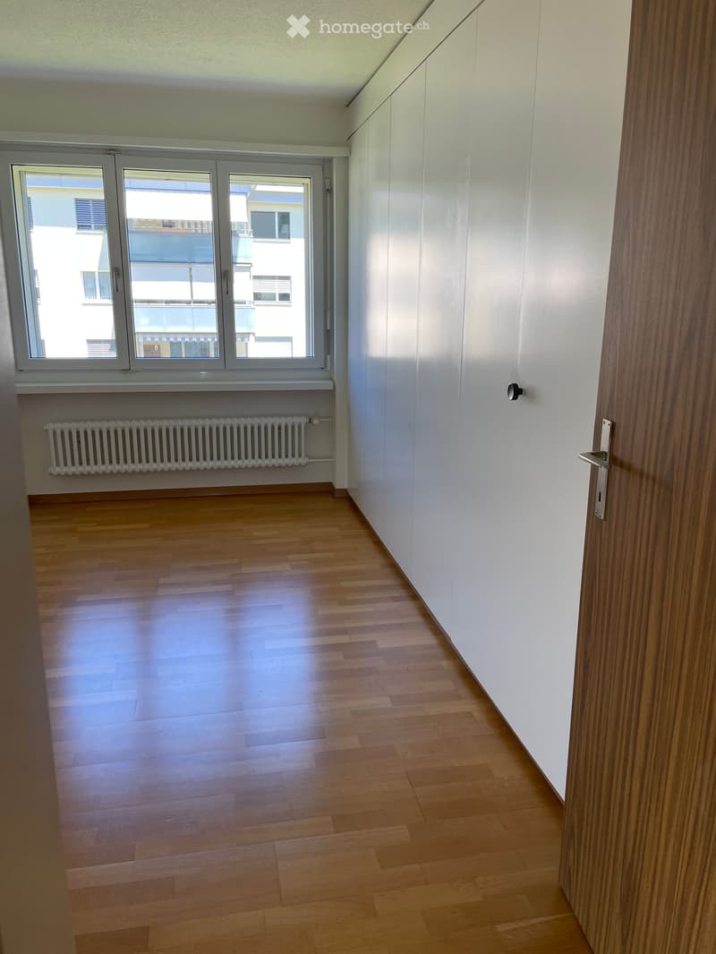 Empty room with wooden flooring, white walls, large window with view, radiator, and a door with a silver handle.