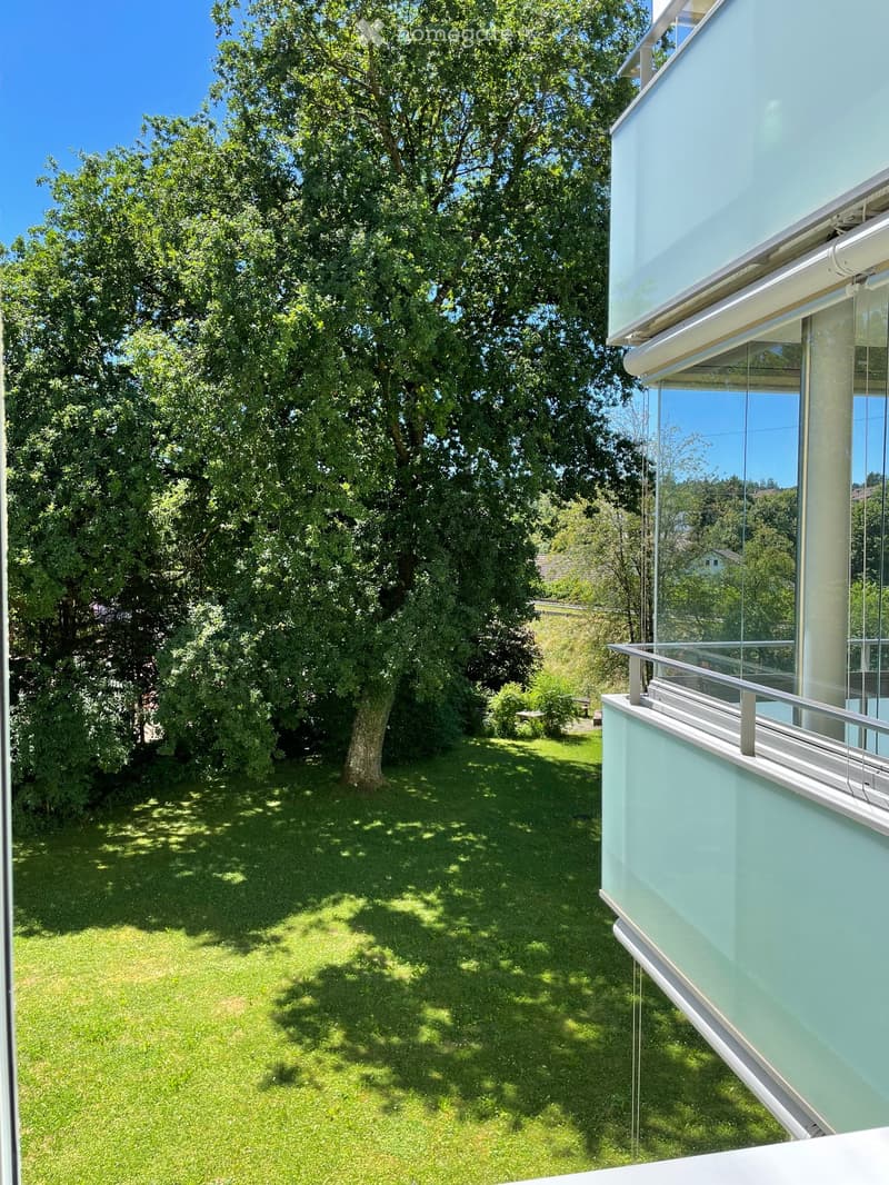 Lush green garden with a large tree, grass, and a modern building with a balcony overlooking the area.