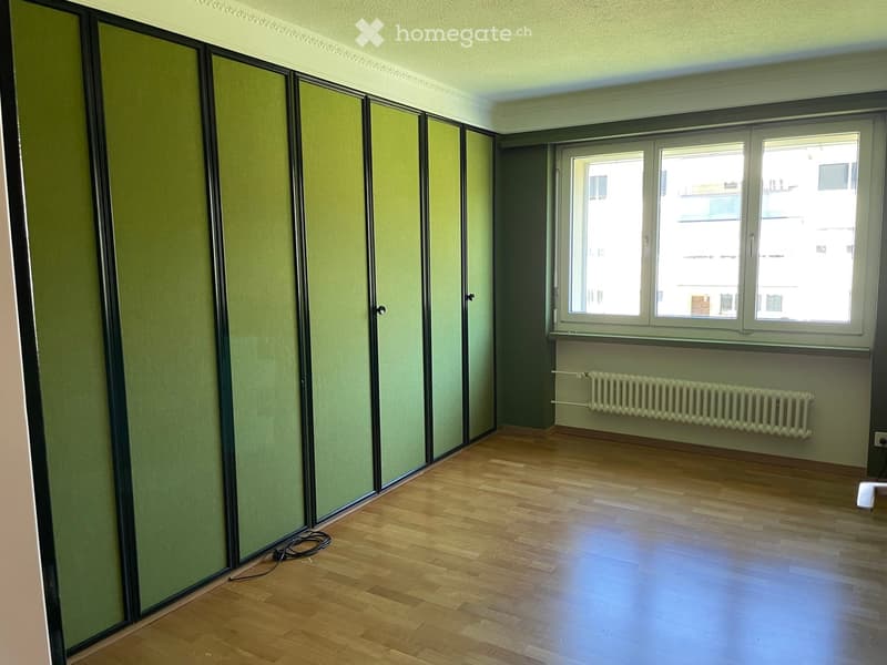Empty room with wooden flooring, green walls, large windows, radiator, and a partially visible cable on the floor.