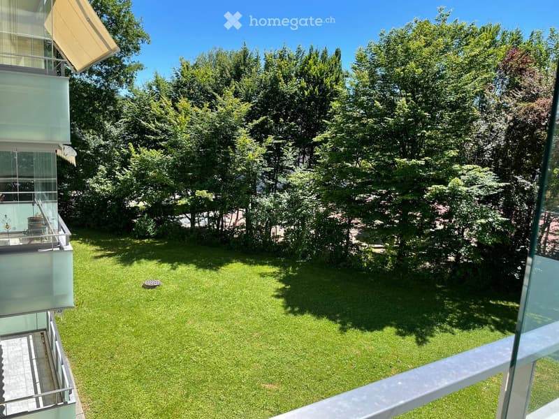 View from a balcony overlooking a well-maintained lawn surrounded by tall trees, with a modern building visible on the left side.