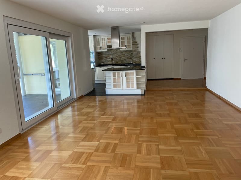 Empty room with polished wooden parquet flooring, large sliding glass door leading to outdoor area, and visible kitchen area in the background.