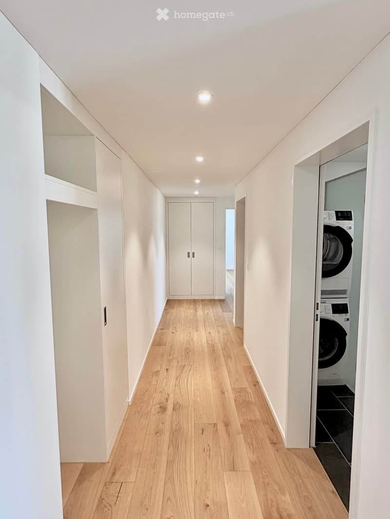 A narrow hallway with light wooden flooring, white walls, and built-in storage cabinets. A washing machine is visible in an adjacent room.