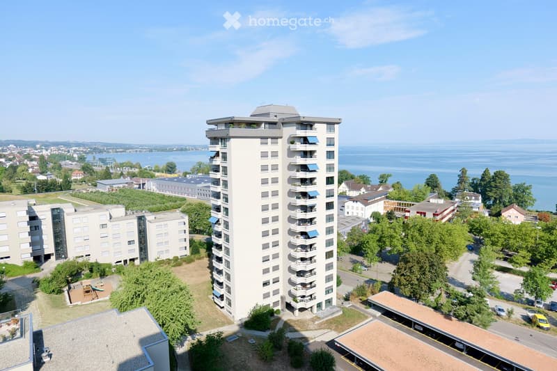A modern multi-story residential building with balconies overlooking a body of water, surrounded by greenery and other structures.