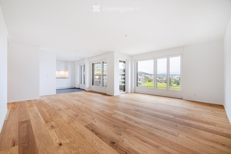 Bright, unfurnished room with wooden flooring, large windows offering a scenic view, and a small kitchen area visible in the background.