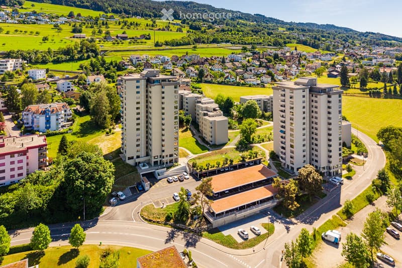Aerial view of a residential complex with multiple high-rise apartment buildings, surrounded by green fields, trees, and smaller houses in the background.