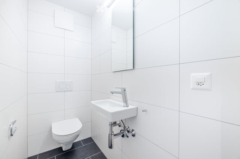 Modern, minimalist bathroom with white tiled walls, a toilet, a sink with a chrome faucet, and a large mirror above the sink.