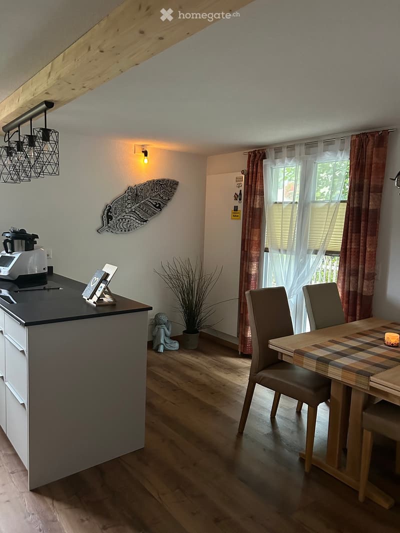 A modern kitchen and dining area with wooden flooring, a grey countertop, a small table with chairs, a potted plant, and decorative items.