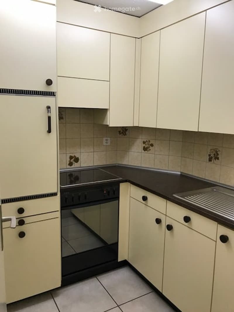 A compact kitchen with white cabinets, a black countertop, and a built-in oven. The space features a stainless steel sink and a refrigerator with black handles.