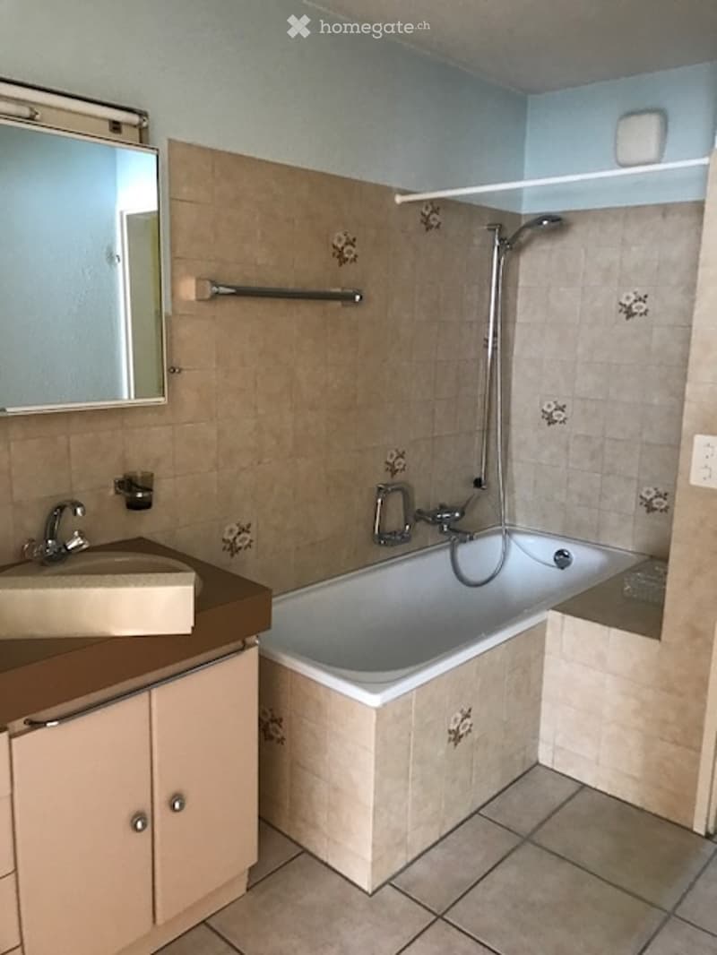 A small bathroom with a bathtub, tiled walls and floor, a mirror above the sink, and a cabinet for storage.