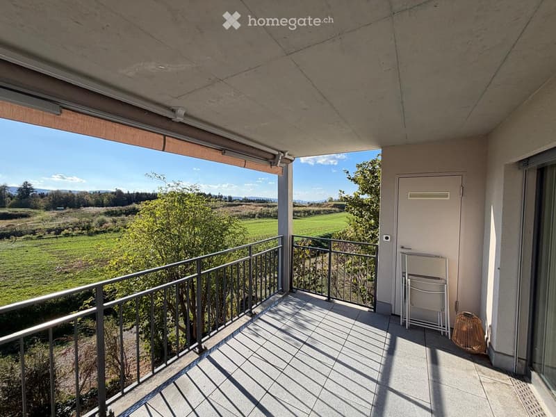 Balcony with tiled floor, metal railing, and a view of a green field and trees. Features a sliding glass door, a white folding chair, and a woven basket.
