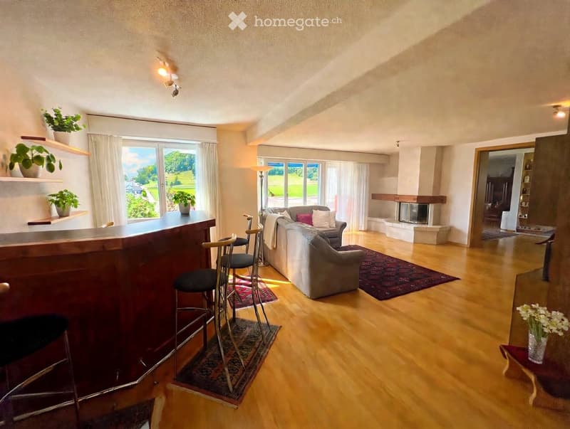 Bright living room with wooden flooring, large windows offering a scenic view, a sofa, chairs, a bar area with stools, and a fireplace.