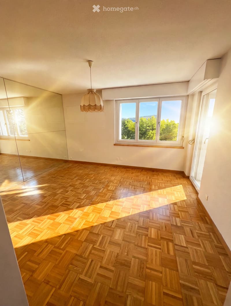 Empty room with wooden herringbone flooring, large windows with a view of greenery, and a hanging light fixture.