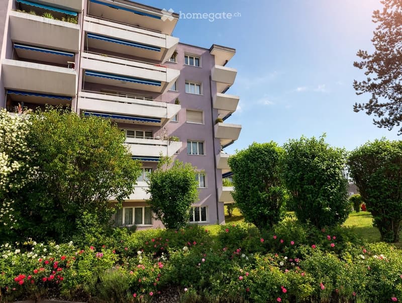 Multi-story apartment building with balconies, surrounded by greenery and flowers.