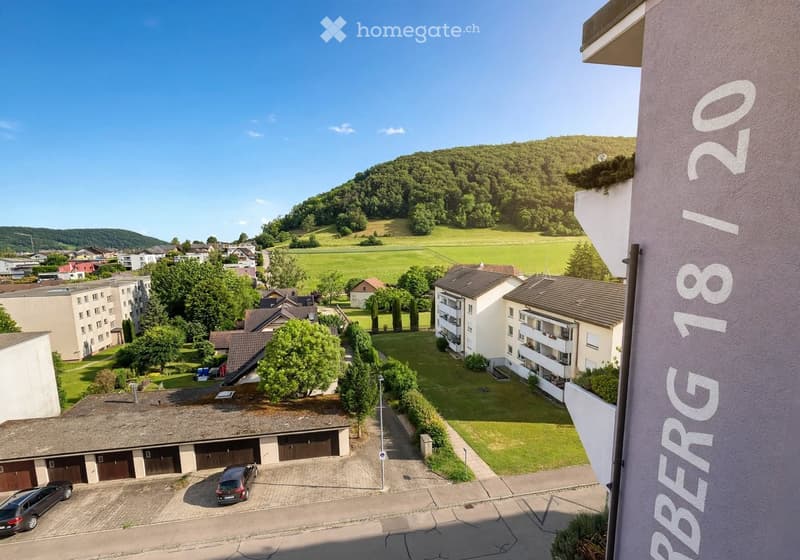 Aerial view of a residential area with apartment buildings, garages, parking spaces, and a scenic view of a green valley and hills in the background.