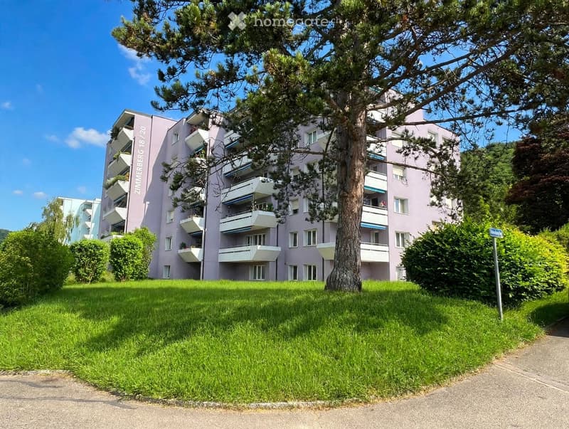 A multi-story residential building with pink facade, balconies, and surrounding greenery.