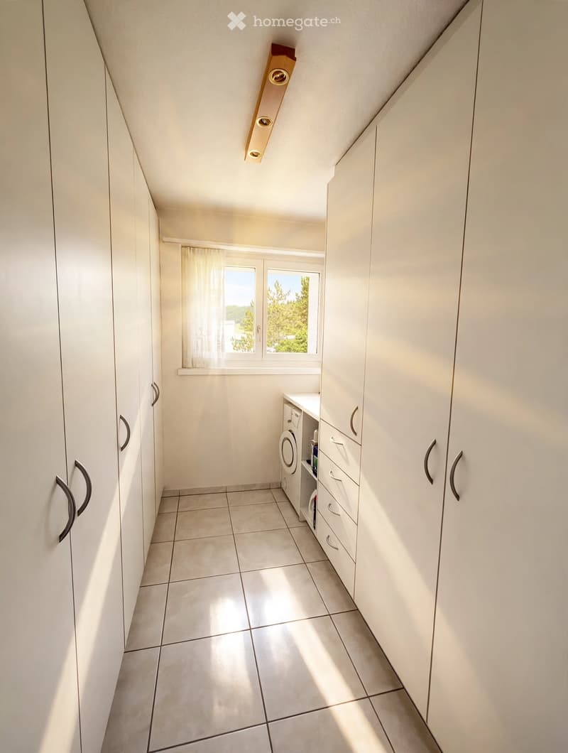 Bright hallway with white tiled floor, white built-in cabinets, and a small window with a view of greenery.