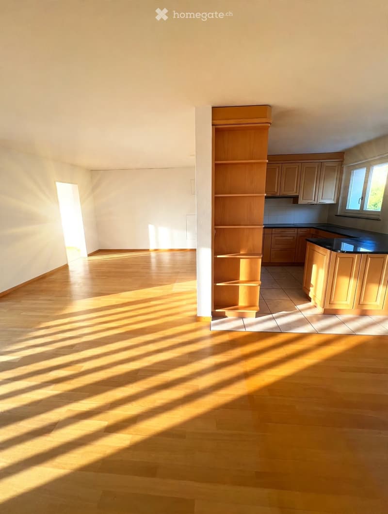 Empty room with wooden flooring and natural light streaming in from a doorway. Adjacent kitchen area with cabinets and countertops is visible.
