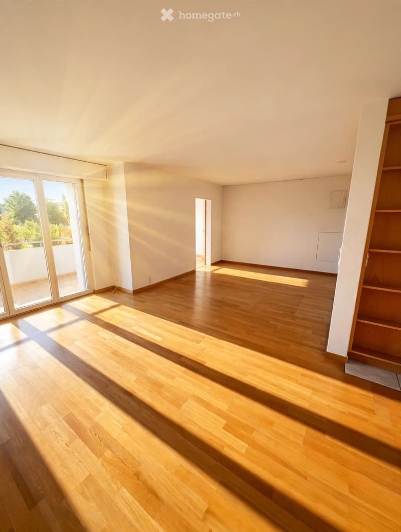 Empty room with wooden flooring, white walls, large sliding glass doors leading to a balcony, and natural light streaming in.