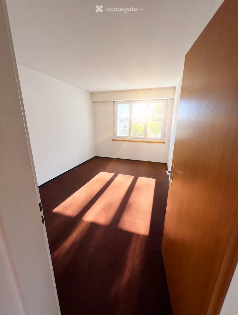 Empty room with brown carpet and white walls, featuring a window with wooden frame and a door with a silver handle.