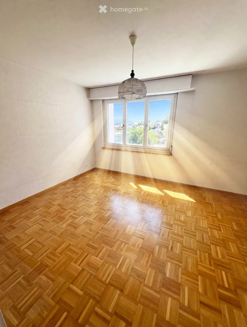 Empty room with wooden herringbone flooring, large windows with a view, and a hanging light fixture.