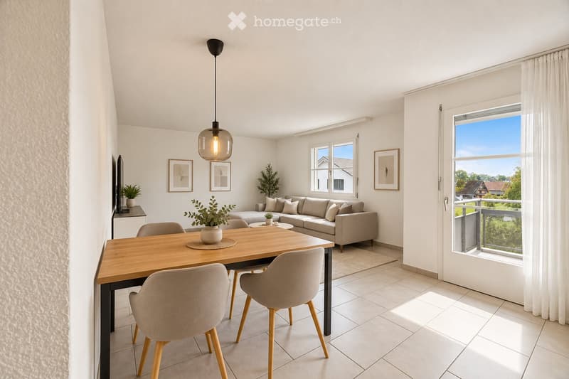 Modern living and dining area with a grey sectional sofa, wooden dining table with four chairs, pendant light, framed pictures, and plants. Large windows and a balcony with a view of the garden.