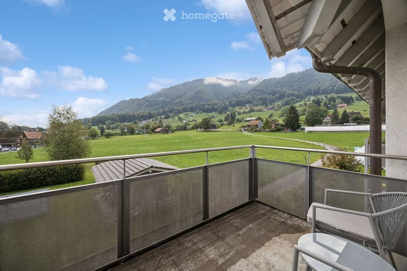 Balcony with a view of a green valley and mountains, featuring a metal railing, a glass table, and a wicker chair.