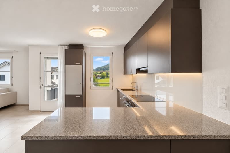 Modern kitchen with dark cabinets, light countertops, stainless steel sink, and a refrigerator. Large windows offer a scenic view of the landscape.