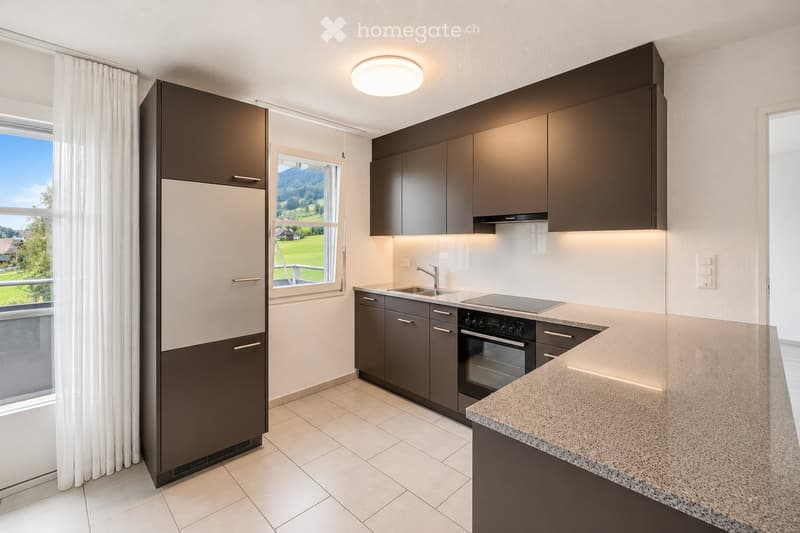 Modern kitchen with dark grey cabinets, a refrigerator, oven, sink, and countertop, featuring large windows with a scenic view.