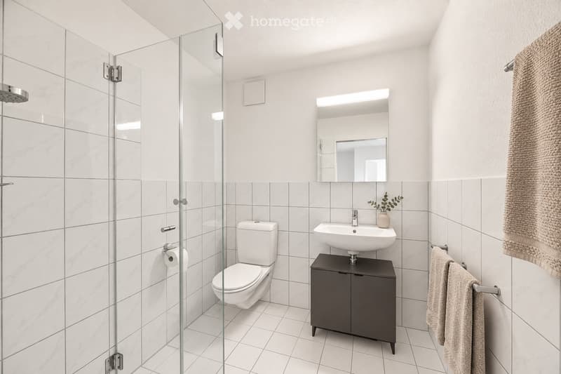 Modern bathroom with white tiles, a glass-enclosed shower, a toilet, a sink with a dark cabinet, and a mirror above the sink. A towel hangs on a rack, and a small plant is placed on the cabinet.