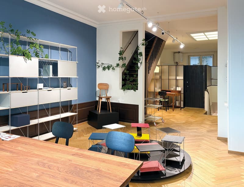 Modern room with white and blue walls, wooden flooring, metal shelving, various colored chairs, a staircase, a wooden desk with a lamp