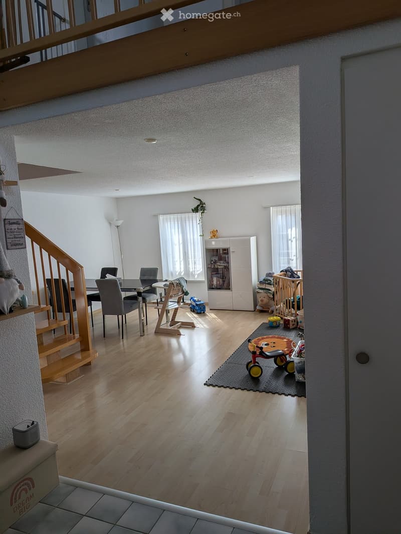 Bright, open living space with wooden flooring, a staircase leading to an upper level, a dining area with chairs and a table, a cabinet, and various children's toys including a tricycle, baby chair, and baby walker.