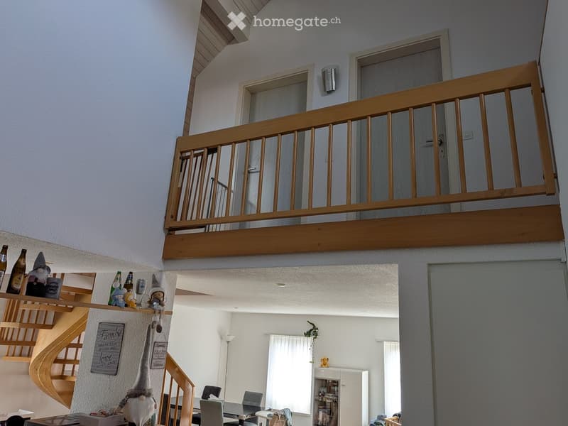 Spacious living area with a dining table, chairs, a cabinet, and decorative gnomes on a shelf. A wooden staircase leads to an upper level with a balcony.