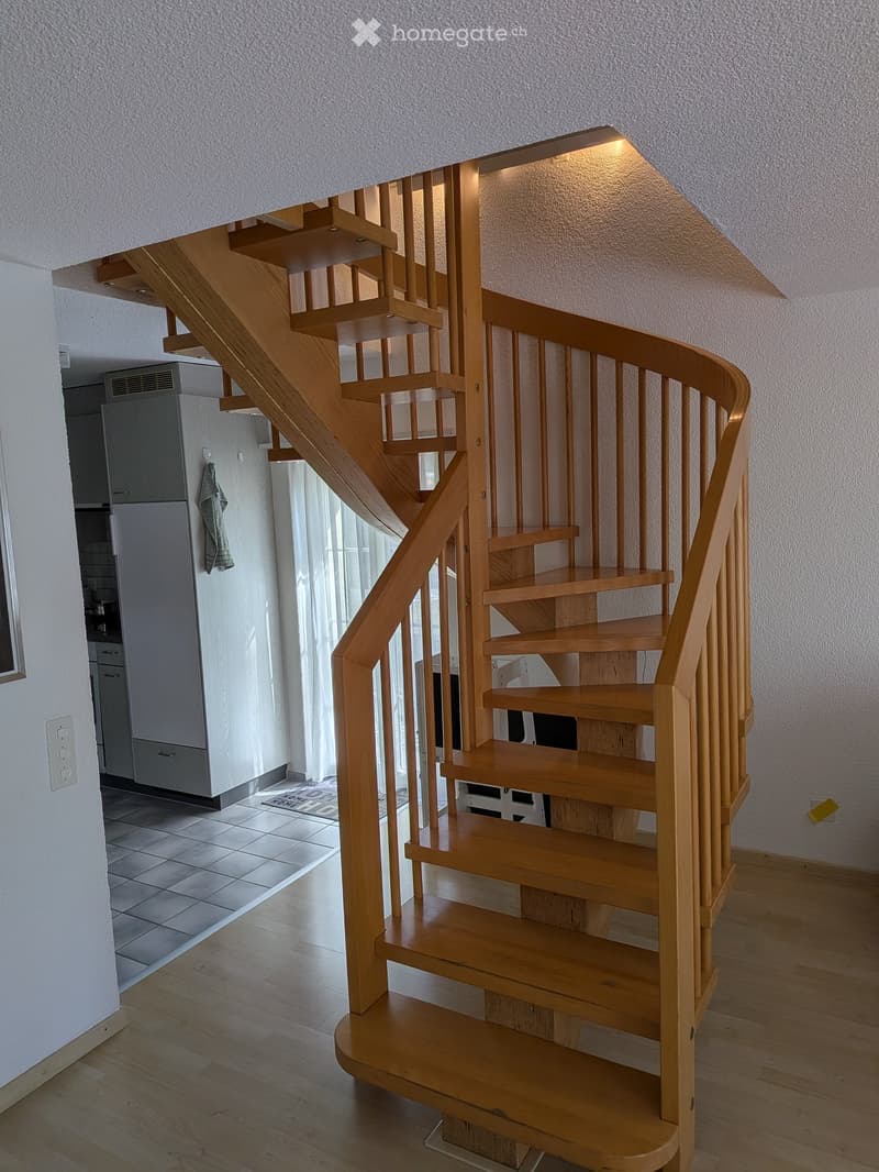 Spiral wooden staircase with wooden handrail and steps, leading to an upper level. The hallway has light wooden flooring and white walls.