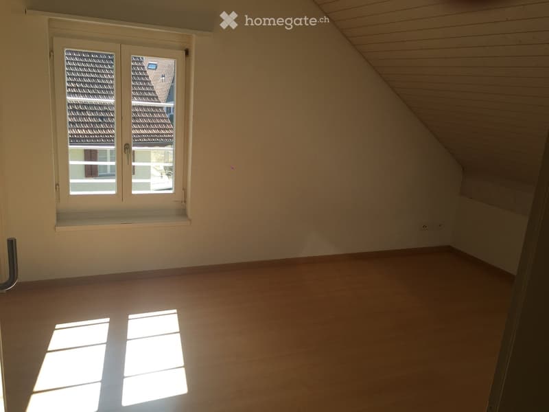 Empty attic room with wooden flooring, white walls, and a large window offering a view of neighboring rooftops.