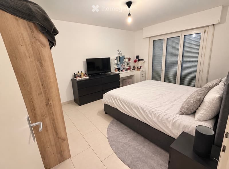 Modern bedroom with a large bed, nightstand, dresser, mirror, and television. The room features large windows with blinds and a sleek, contemporary design.