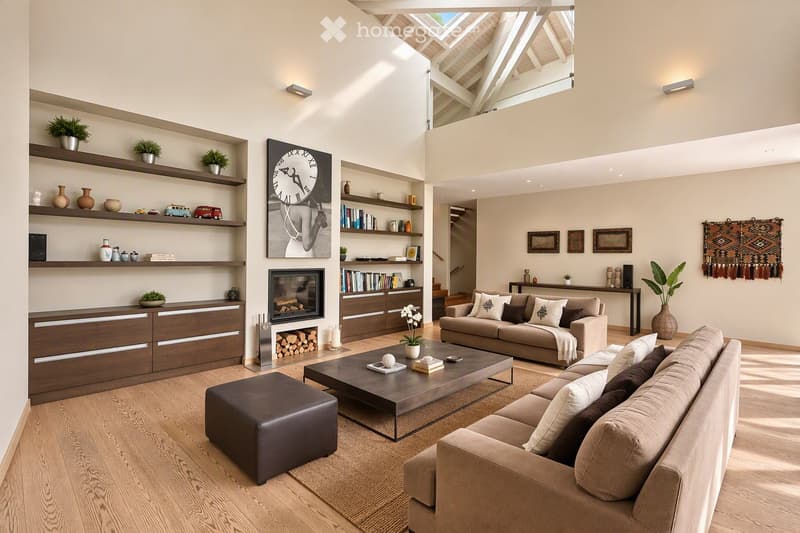 Modern living room with light-colored walls, wooden flooring, and large windows. Features include a fireplace, two sofas, a coffee table, an ottoman, built-in bookshelves, decorative vases, plants, and artwork.